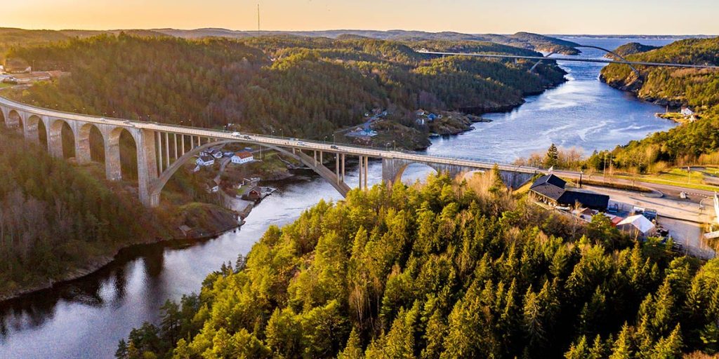 Gamla Svinesundsbron från ovan, i varmt eftermiddagsljus. Himlen och vattnet är matchande blå och grönskan frodar på omgivande träd.